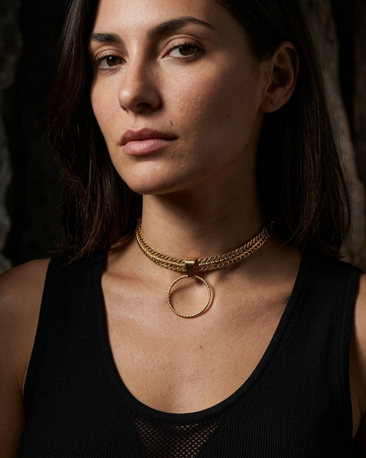 A close-up of a woman wearing a gold multi-strand chain choker necklace with a large circular ring pendant, paired with a black ribbed tank top.