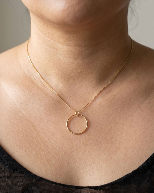 A close-up of a person's neck and upper chest wearing a delicate gold chain necklace with a thin circular pendant, paired with a black top.