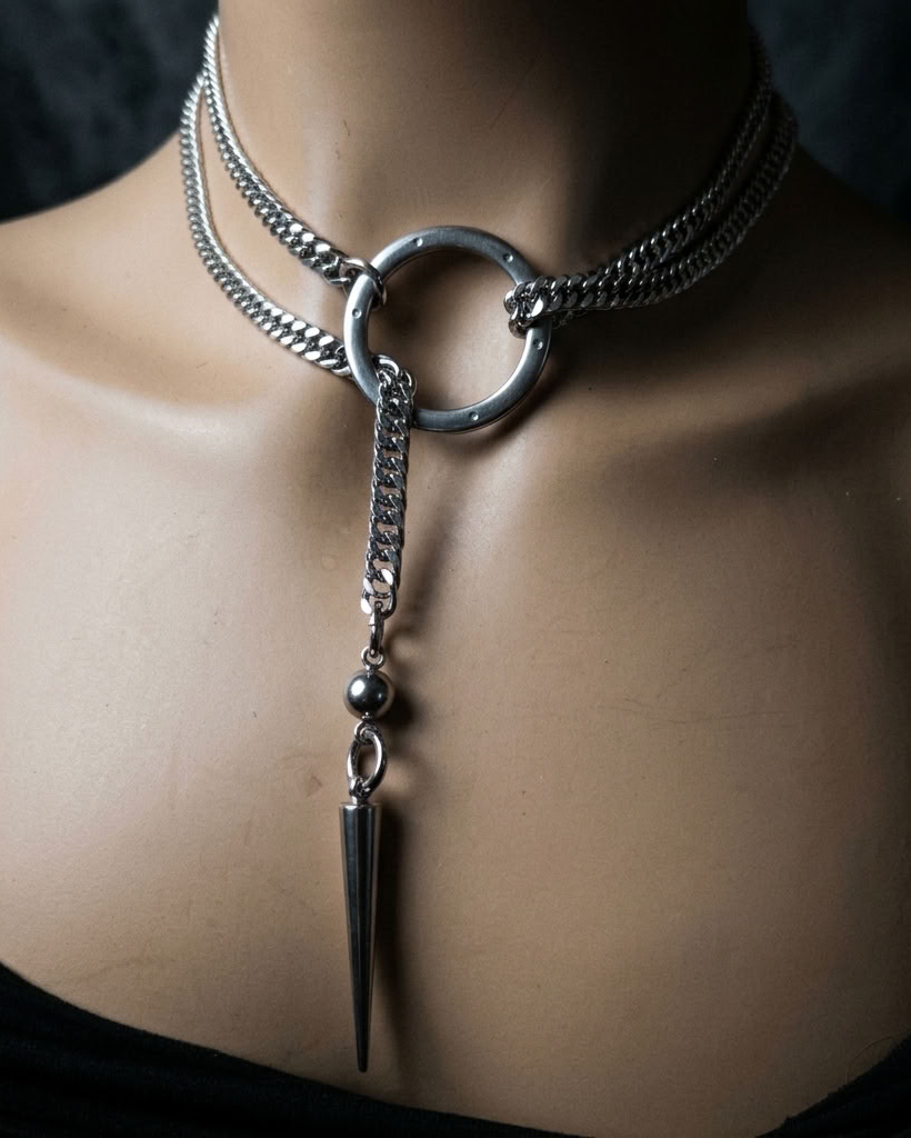 A silver-toned gothic choker necklace displayed on a mannequin torso. The necklace features a thick chain layered with a thinner chain, a large central ring, a dangling chain with a small bead, and a long pointed spike charm.