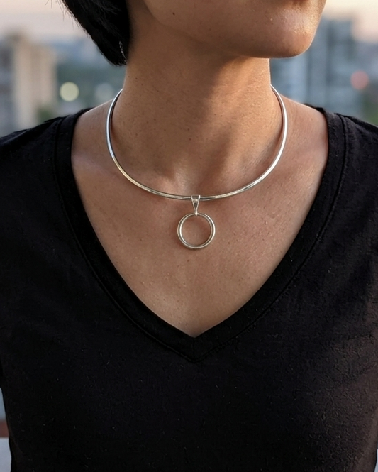 A woman wearing a minimalist silver necklace with a circular pendant, photographed from the chest up against a softly blurred city skyline at dusk.