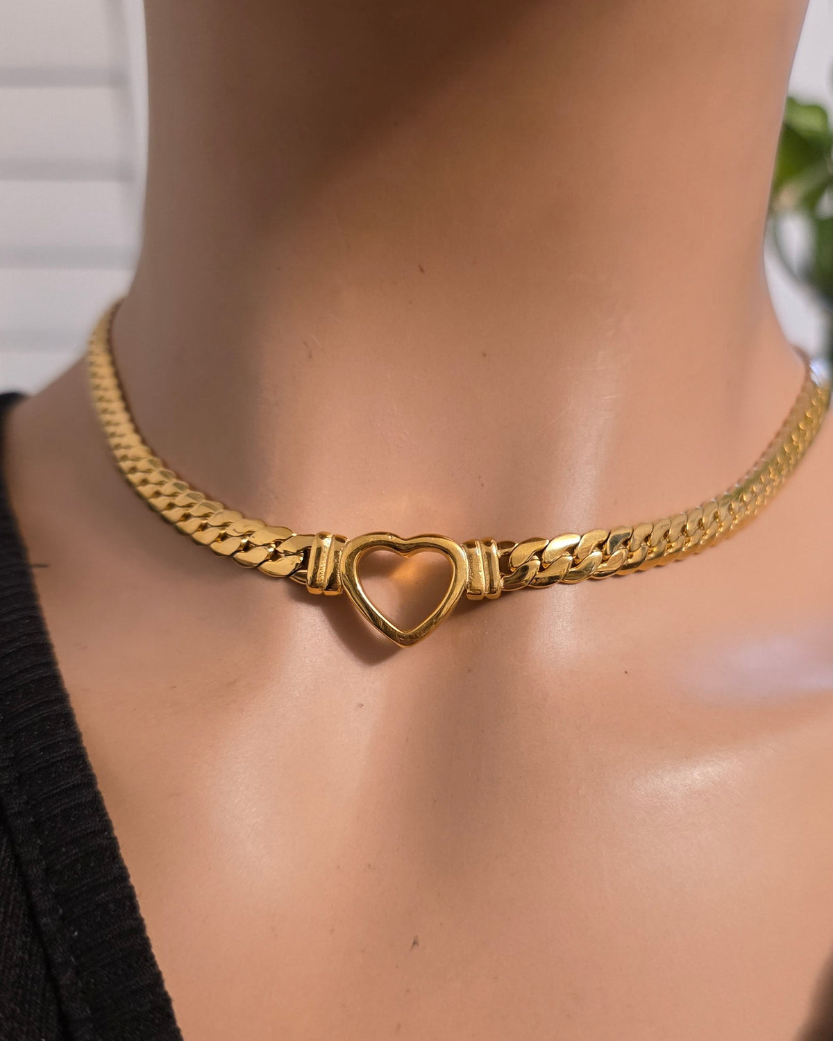 A gold chain choker necklace with a heart-shaped clasp, displayed on a mannequin bust wearing a black top.