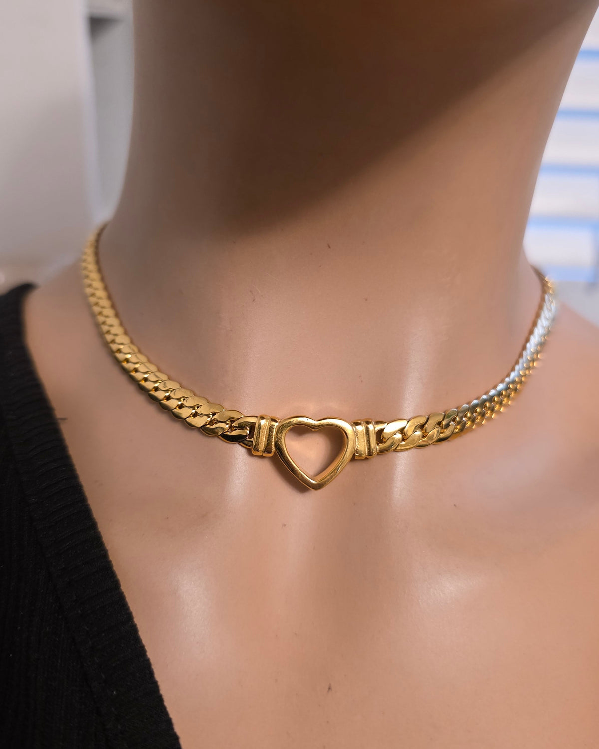 A gold chain choker necklace with a heart-shaped clasp, displayed on a mannequin neck wearing a black top.