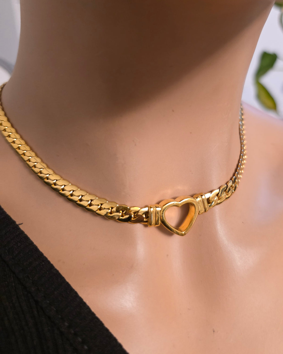A close-up of a gold chain necklace with a heart-shaped clasp, displayed on a mannequin neck wearing a black top.