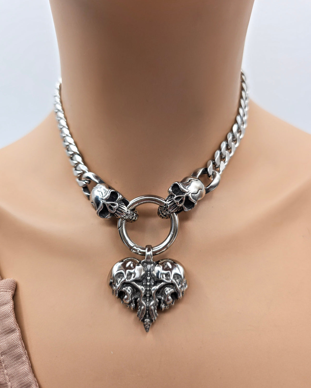 A silver-toned metal choker necklace displayed on a mannequin neck. The necklace features a chunky curb chain, skull accents on both sides of a central ring, and a large ornate heart-shaped pendant with a skull motif.