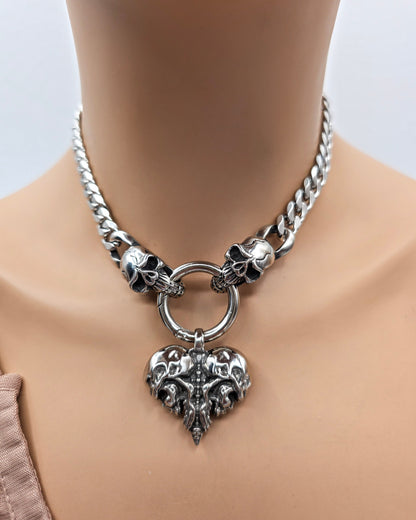 A silver-toned metal choker necklace displayed on a mannequin neck. The necklace features a chunky curb chain, skull accents on both sides of a central ring, and a large ornate heart-shaped pendant with a skull motif.