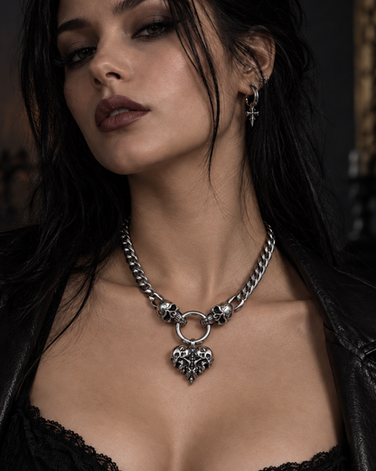 A close-up of a woman wearing a chunky silver chain necklace with a heart-shaped pendant, styled with a black leather jacket and black lace top in a dimly lit gothic setting.
