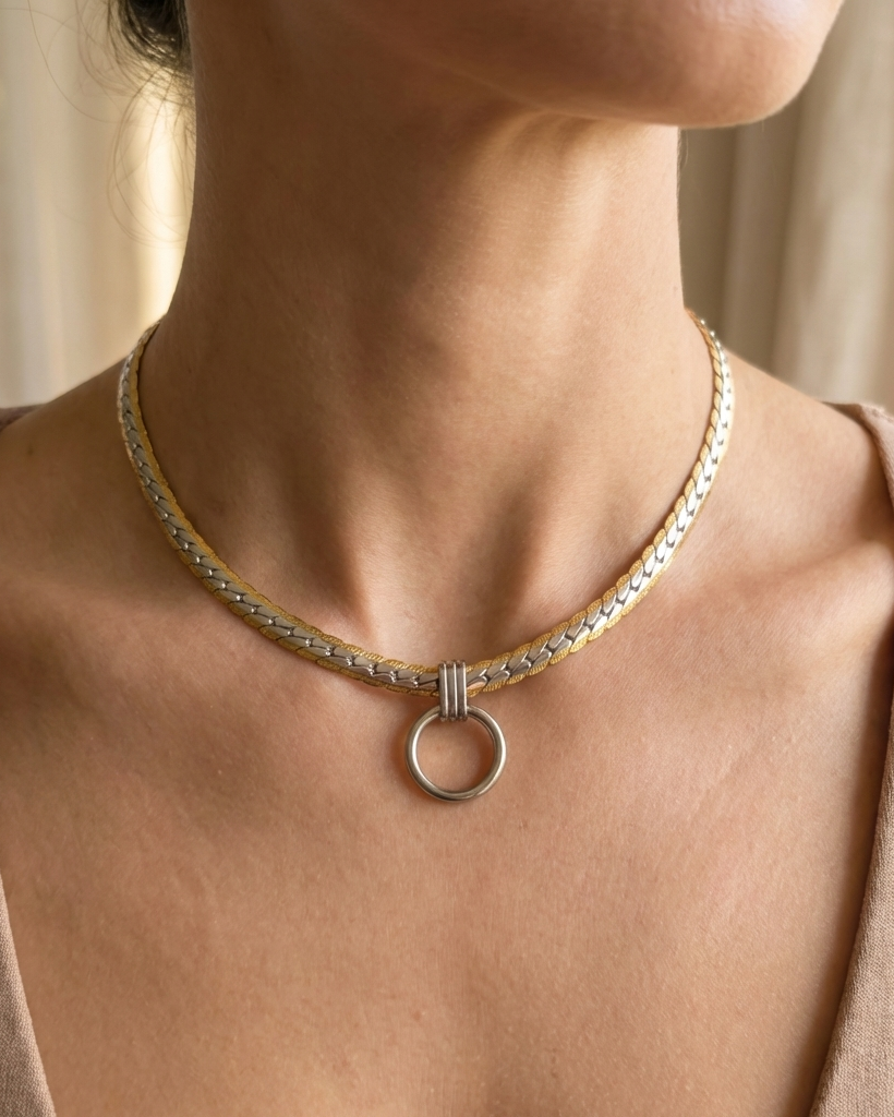 A close-up of a person's neck and upper chest wearing a delicate chain necklace with a silver-toned circular ring pendant and a small gold-toned accent.