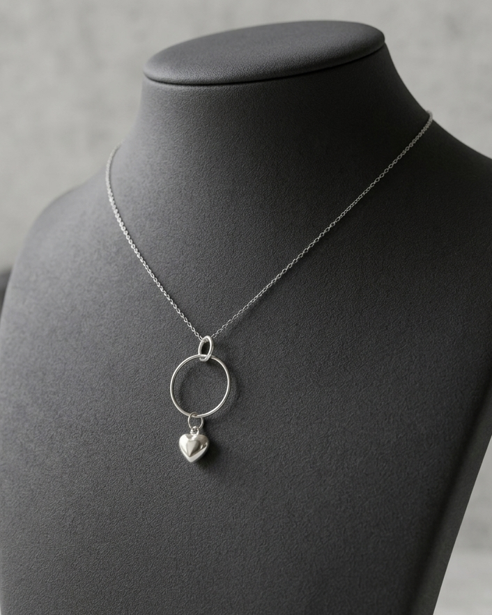A silver necklace with a large open circle pendant and a small dangling heart charm, displayed on a black jewelry bust against a soft gray background.