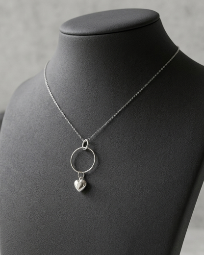 A silver necklace with a large open circle pendant and a small dangling heart charm, displayed on a black jewelry bust against a soft gray background.