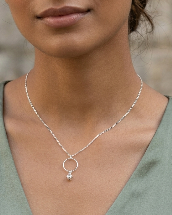 A woman wearing a delicate silver necklace with a small geometric pendant, photographed in a close-up portrait against a softly blurred stone wall.