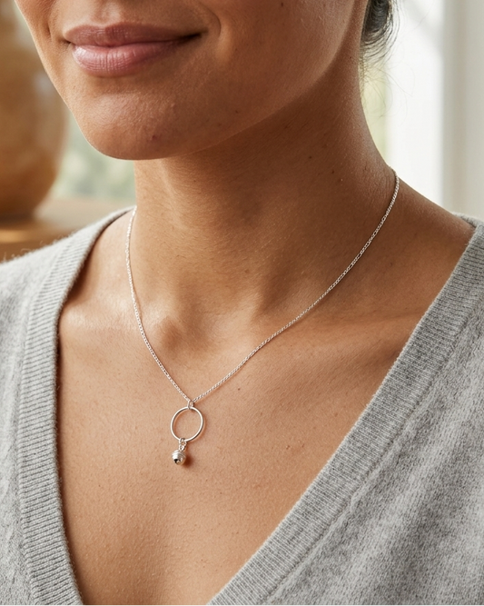 A close-up lifestyle shot of a woman wearing a delicate silver necklace with a minimalist ring and teardrop pendant, styled with a gray knit sweater in a softly lit indoor setting.
