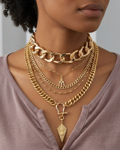 A close-up of a model wearing a layered gold necklace set, including a chunky chain choker, a curb chain, a rope chain, and a delicate chain with a small pendant and a larger diamond-shaped pendant, paired with a mauve-colored top.