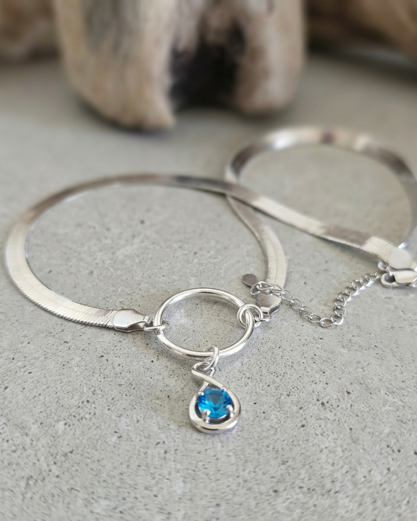 A silver-toned snake chain bracelet with a circular ring, a small chain extender, and a dangling teardrop-shaped blue gemstone, displayed on a textured gray surface.