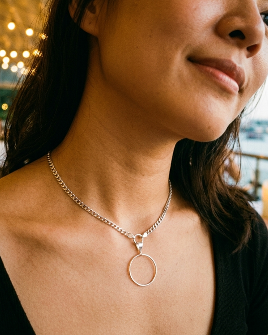 A close-up lifestyle shot of a woman wearing a delicate silver chain necklace with a minimalist circular pendant, set against a warm, softly blurred background with lights and water.