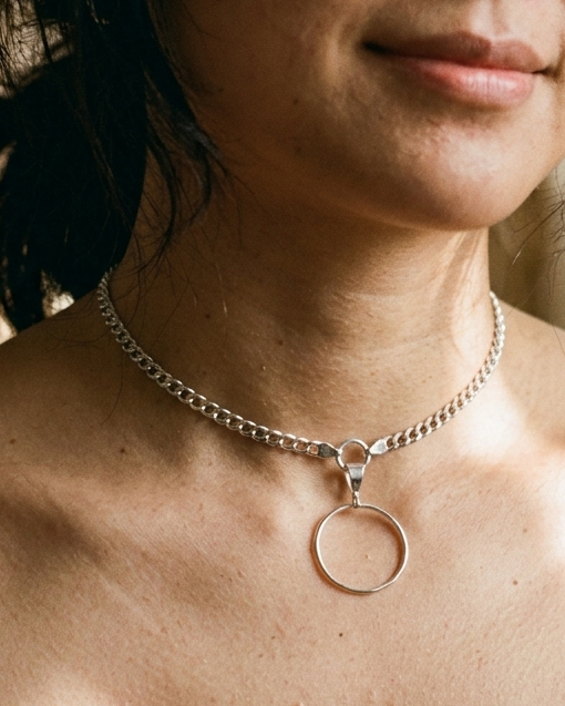 A close-up lifestyle shot of a woman wearing a silver chain choker necklace with a minimalist circular pendant, set against a warm, softly blurred indoor background with candlelight.