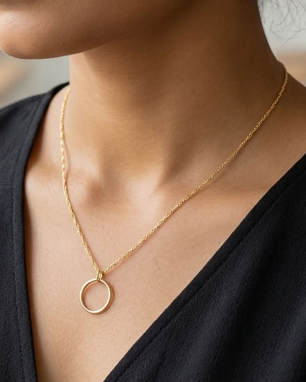 A woman wearing a delicate gold chain necklace with a minimalist open circle pendant, styled with a black V-neck top in a softly blurred indoor setting.