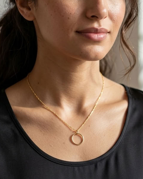 A woman with dark hair tied back wears a delicate gold necklace with a small circular pendant, paired with a black scoop-neck top. She is photographed in a softly lit indoor setting with a blurred background showing shelves and a plant.