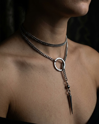 A close-up of a model's neck and upper chest wearing a layered silver-toned necklace with a choker chain, a longer chain featuring a circular ring pendant, and a dangling spike charm.