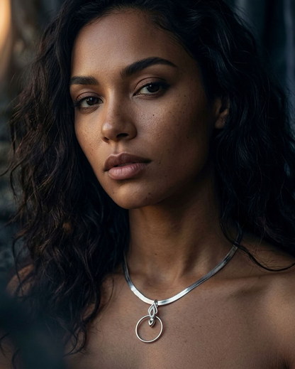 A close-up portrait of a woman with dark curly hair wearing a polished silver necklace with a double-ring pendant, set against a dark, moody background.