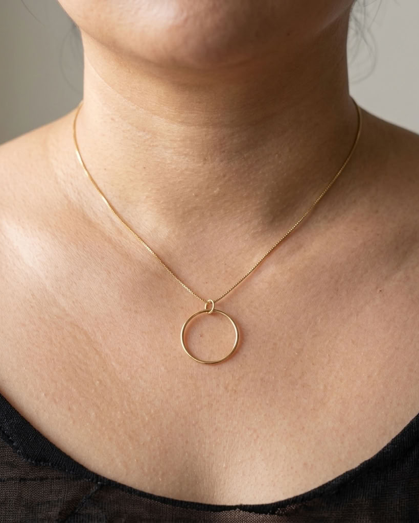 A close-up of a person's neck and upper chest wearing a delicate gold chain necklace with a thin circular pendant, paired with a black top.