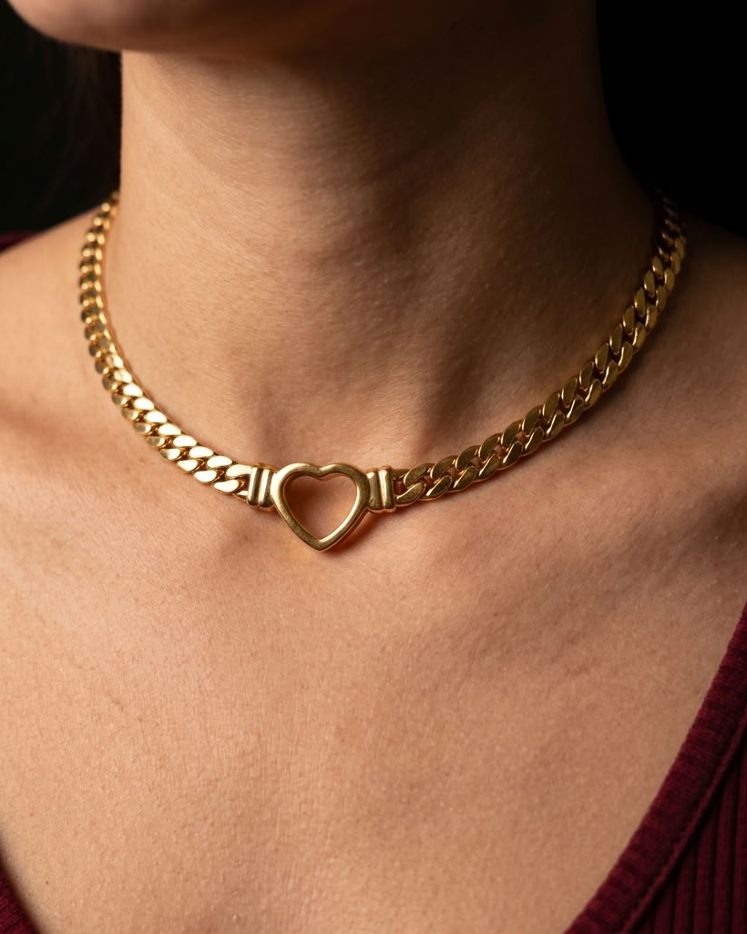 A close-up of a gold chain necklace with a heart-shaped center clasp, worn by a person in a burgundy ribbed top against a dark background.