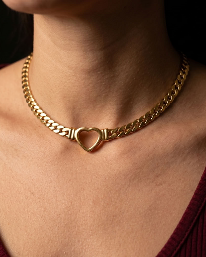 A close-up of a gold chain necklace with a heart-shaped center clasp, worn by a person in a burgundy ribbed top against a dark background.
