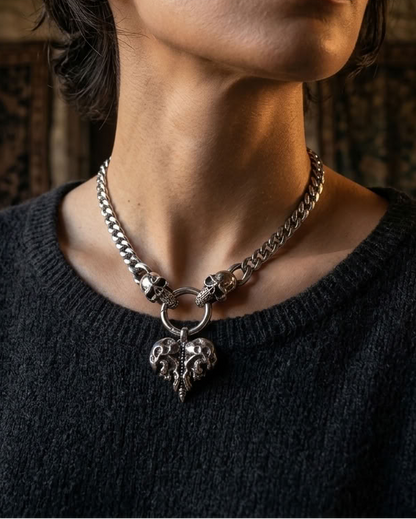 A close-up of a woman wearing a silver chain necklace with a skull ring and skull pendant, paired with a dark gray knit sweater in a dimly lit indoor setting.