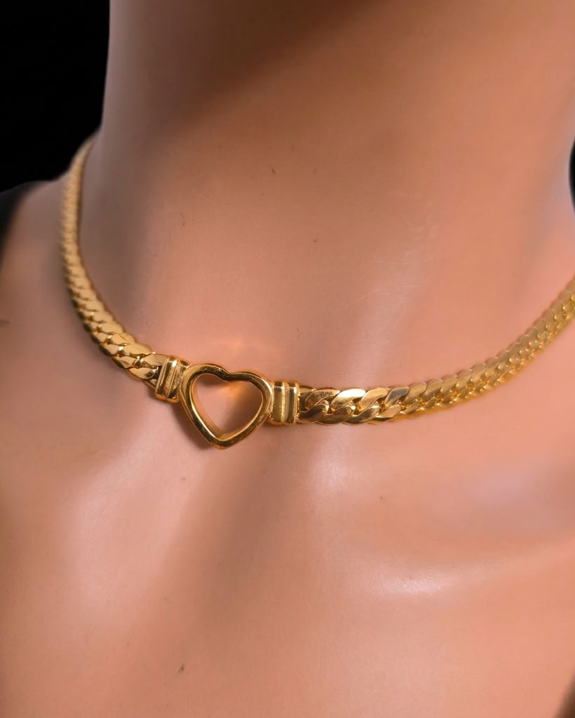 A gold chain necklace with a heart-shaped clasp, shown on a mannequin bust wearing a black top against a dark background.