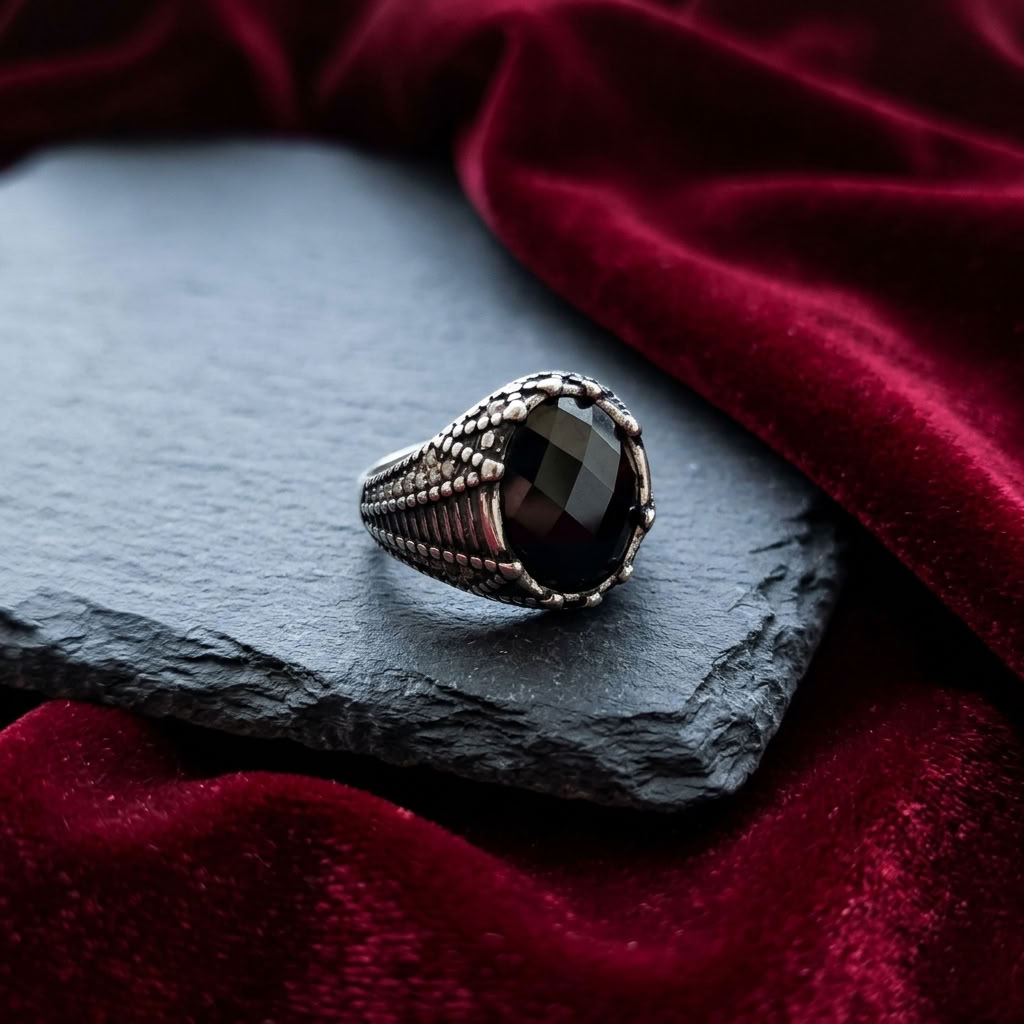 A gothic-style silver ring with a large oval black faceted gemstone, resting on a dark slate slab surrounded by deep red velvet fabric.