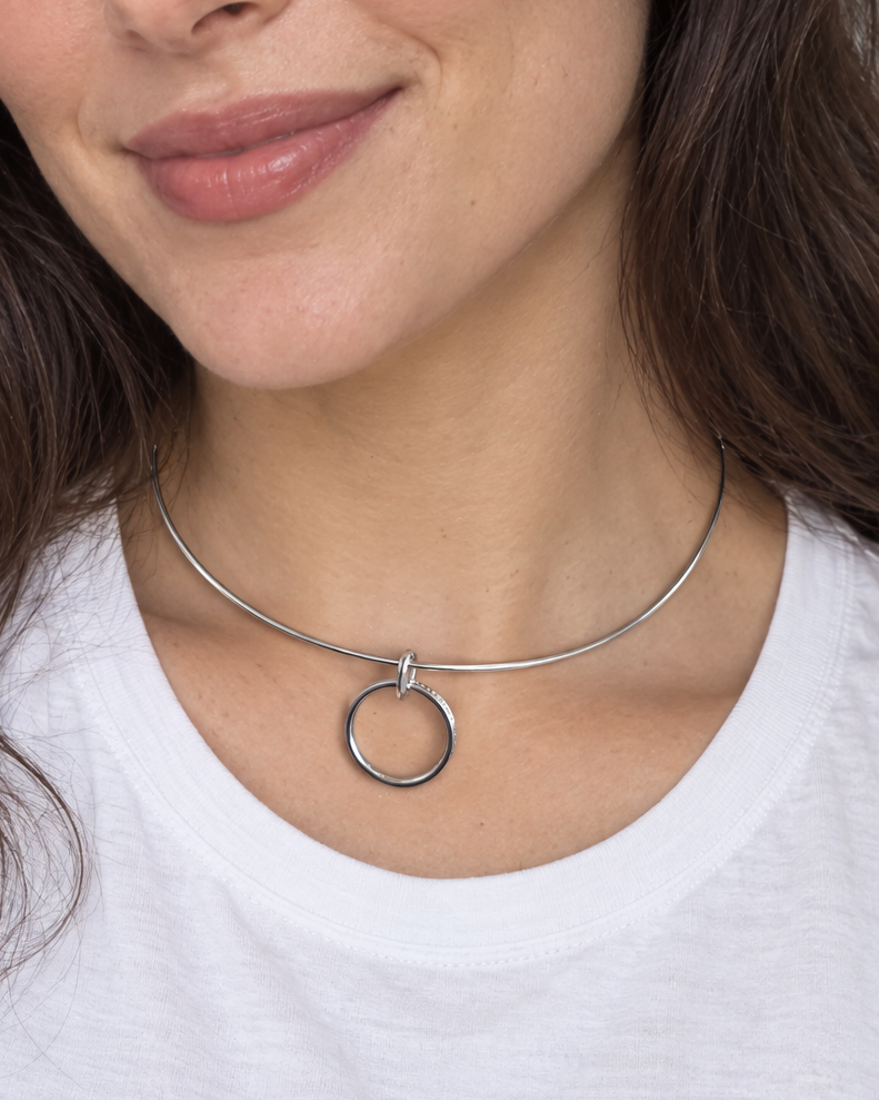 A woman with long brown hair wearing a white t-shirt and a thin silver choker necklace with a circular ring pendant, photographed in a brightly lit indoor setting.