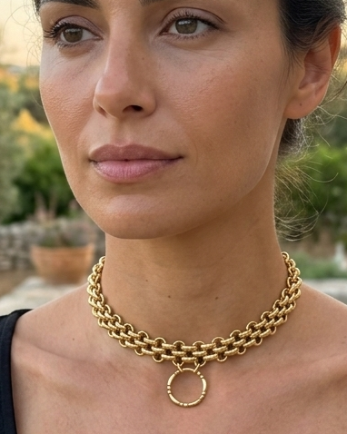 A woman wearing a gold chain necklace with a circular ring pendant, styled with a black scoop-neck t-shirt, photographed outdoors against a stone wall and olive trees at sunset.