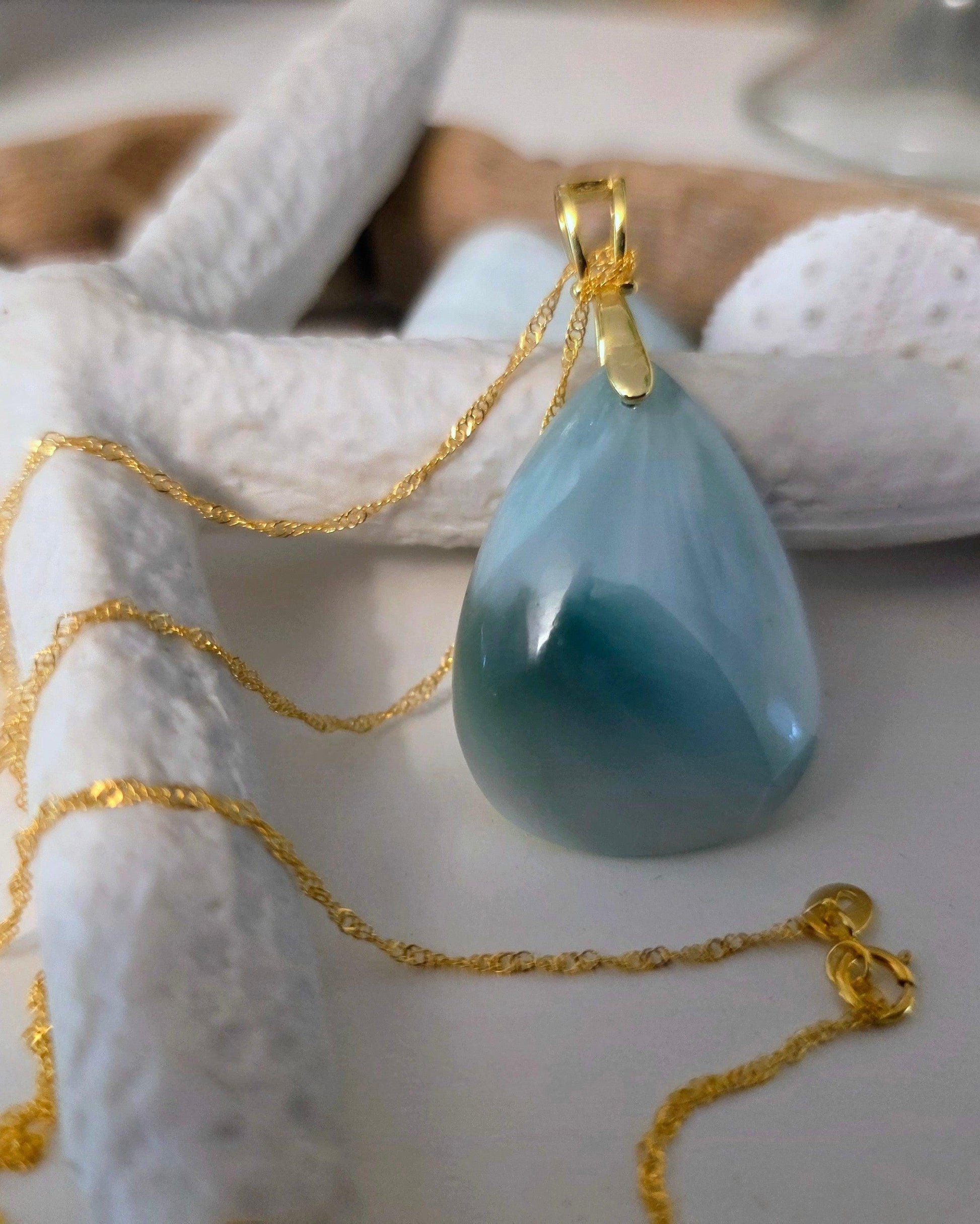 Teardrop-shaped blue pendant on a gold chain with starfish and shell in the background
