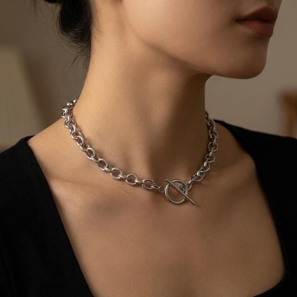 May include: A silver-colored chain necklace with large, rounded links. The necklace features a toggle clasp closure. The jewelry is worn around the neck, and the background is blurred.