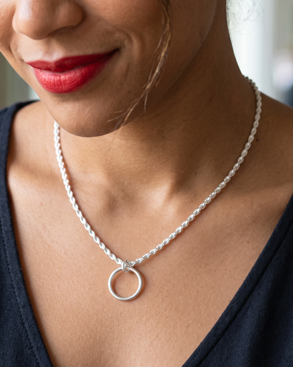 A close-up lifestyle shot of a woman wearing a delicate silver chain necklace with a minimalist circular pendant, set against a soft-focus indoor background.