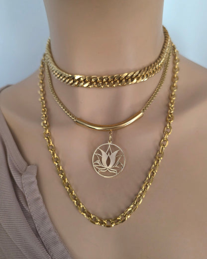 A close-up of a mannequin neck wearing three layered gold necklaces: a chunky curb chain choker, a delicate chain with a curved bar pendant, and a rope-style chain with a circular pendant featuring a lotus design.