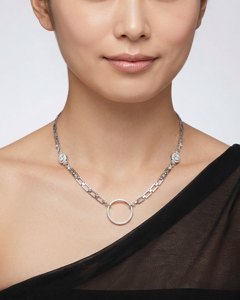 A close-up of a model wearing a silver-toned chain necklace with a central textured circular pendant and small floral details on the chain, paired with a black one-shoulder top against a light gray background.