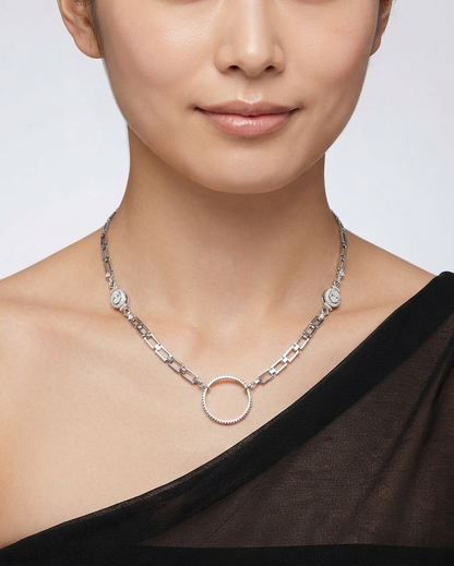 A close-up of a model wearing a silver-toned chain necklace with a central textured circular pendant and small floral details on the chain, paired with a black one-shoulder top against a light gray background.