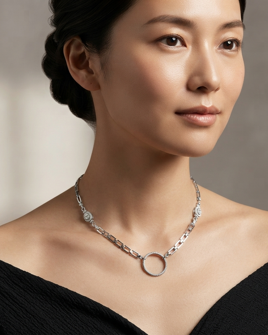 A close-up of a woman wearing a silver chain necklace with a circular pendant and floral details, paired with a black off-the-shoulder top against a soft gray background.