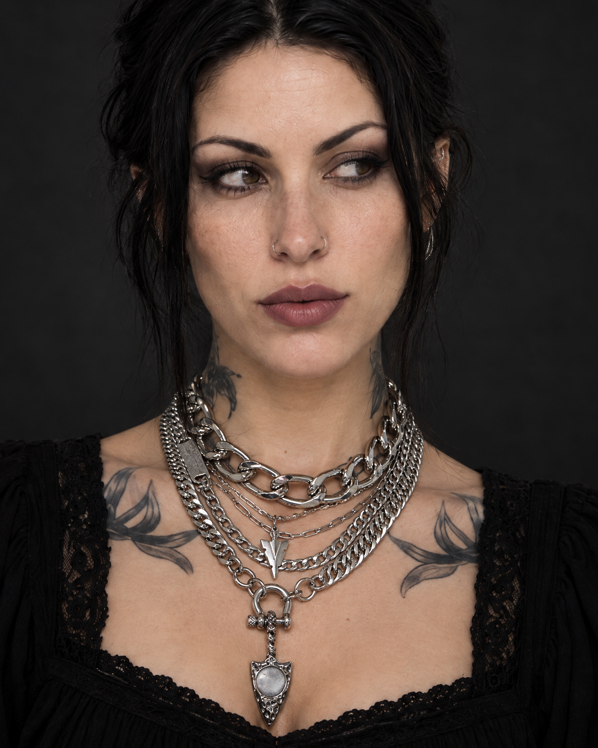A close-up portrait of a woman with dark hair and tattoos wearing multiple layered silver chain necklaces, including a prominent pendant necklace, against a dark studio background.