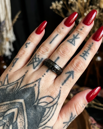 Hand with red nails and tattoos wearing a black ring, with a blurred natural background.